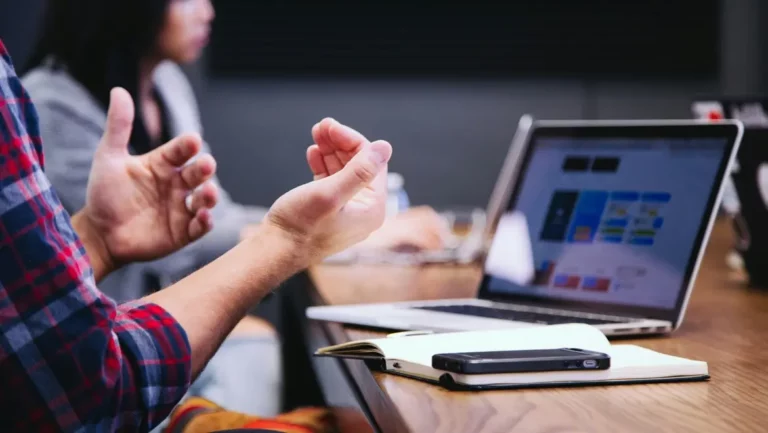 Person gesturing during a collaborative discussion about UI/UX design, with a laptop displaying design elements and a notebook on the table, reflecting Goran Djuran's focus on creating engaging digital experiences.